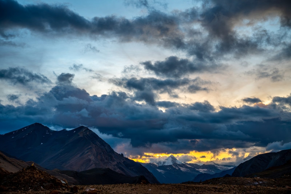 Dark storm clouds over Himalayan peaks with a golden sunset glowing behind a snow-capped summit