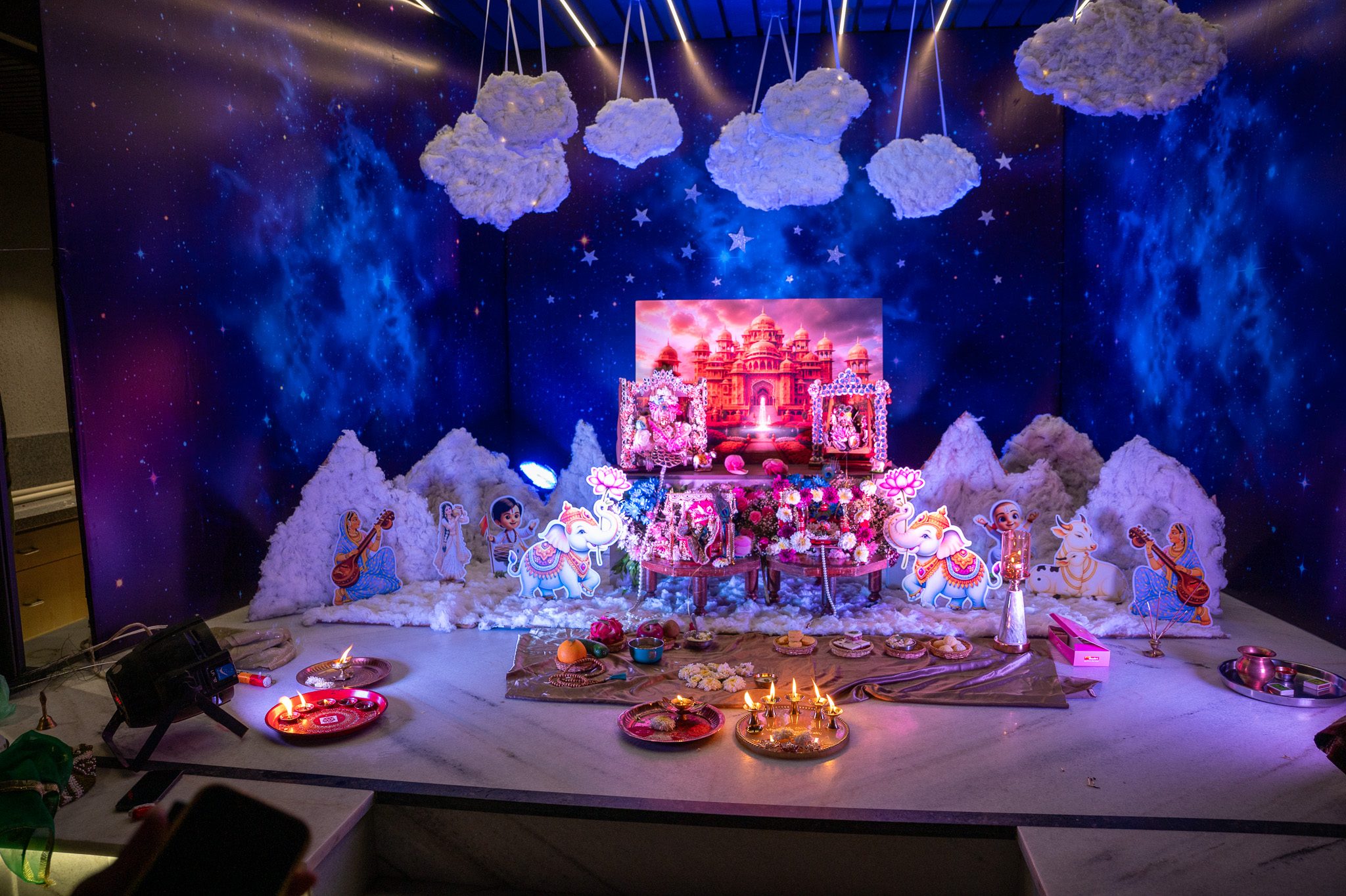 Janmashtami decoration at home with cotton-cloud mountains, cutouts of gopis and elephants, and a Laddu Gopal jhula against a Vrindavan palace backdrop
